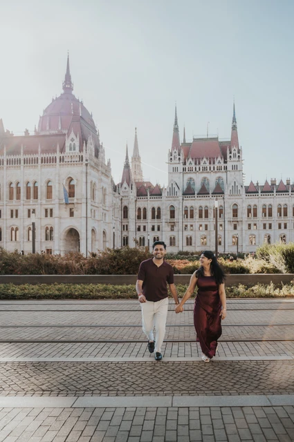 Walking beside Parliament’s flowerbeds at a Budapest couple photoshoot The couple walking hand in hand along the paving beside Parliament’s flowerbeds during their couple photoshoot in Budapest.