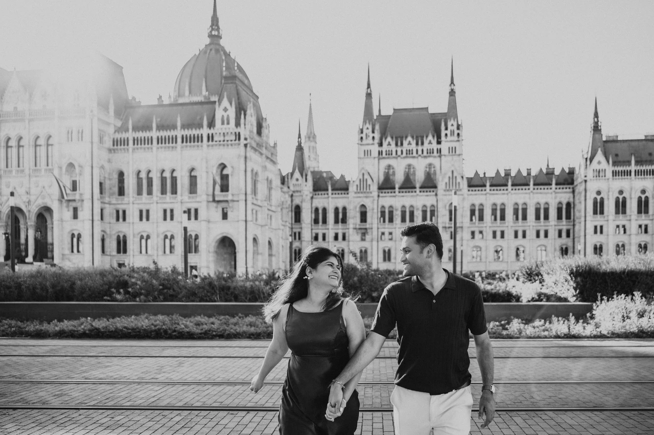 A Budapest couple photoshoot walk in black and white Black-and-white landscape portrait of the couple smiling at each other while walking beside Parliament during their couple photoshoot in Budapest.