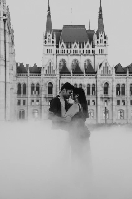 Black-and-white portrait of the couple leaning close through a soft veil of mist, with Parliament behind them, during their couple photoshoot in Budapest.