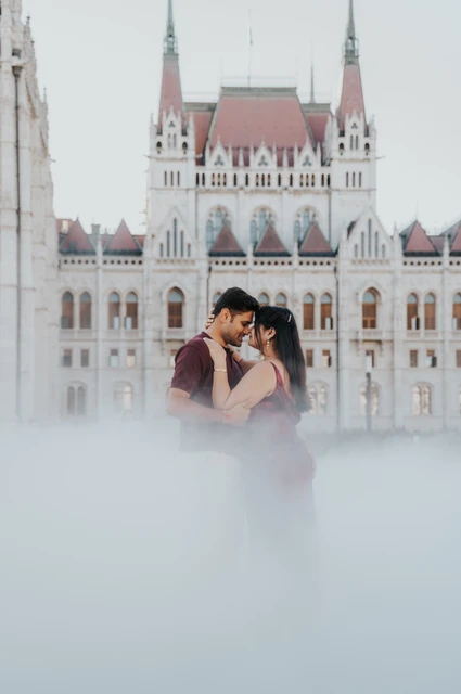 A Budapest couple photoshoot in the mist beside Parliament The couple leaning close with Parliament’s Gothic façade behind them and mist in the foreground during their Budapest couple photoshoot.