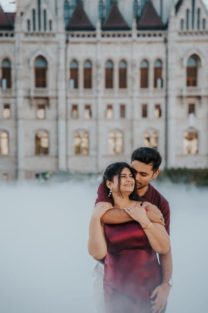 An embrace through the mist at a Budapest couple photoshoot The man embracing his partner from behind in front of Parliament’s arched windows, with mist below them, during their Budapest couple photoshoot.
