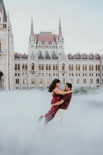 A playful lift beside Parliament at a Budapest couple photoshoot The man lifting his partner in front of Parliament, with soft mist across the foreground, during their playful couple photoshoot in Budapest.