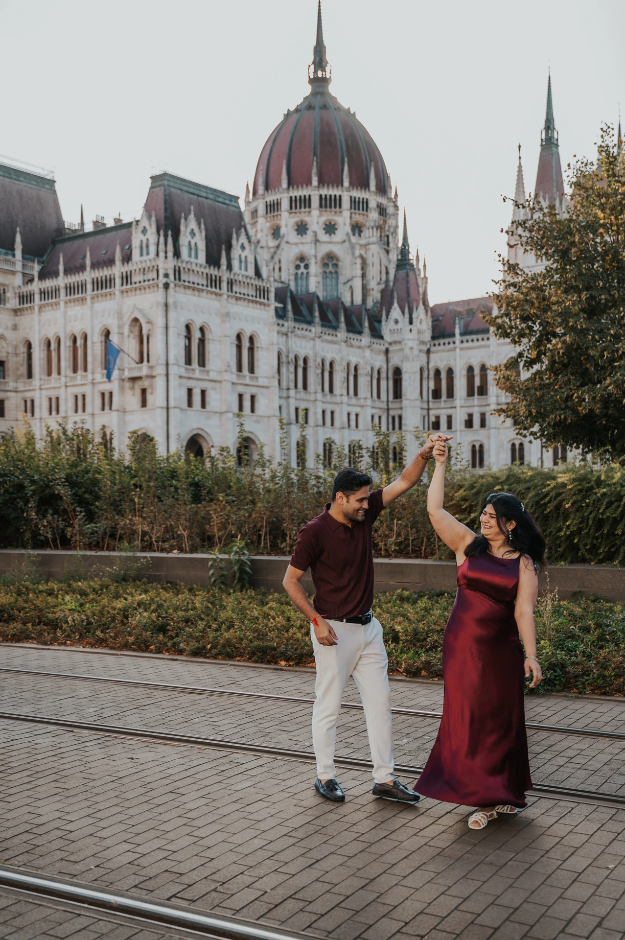 The couple raising their joined hands for a playful turn beside Parliament during their couple photoshoot in Budapest.