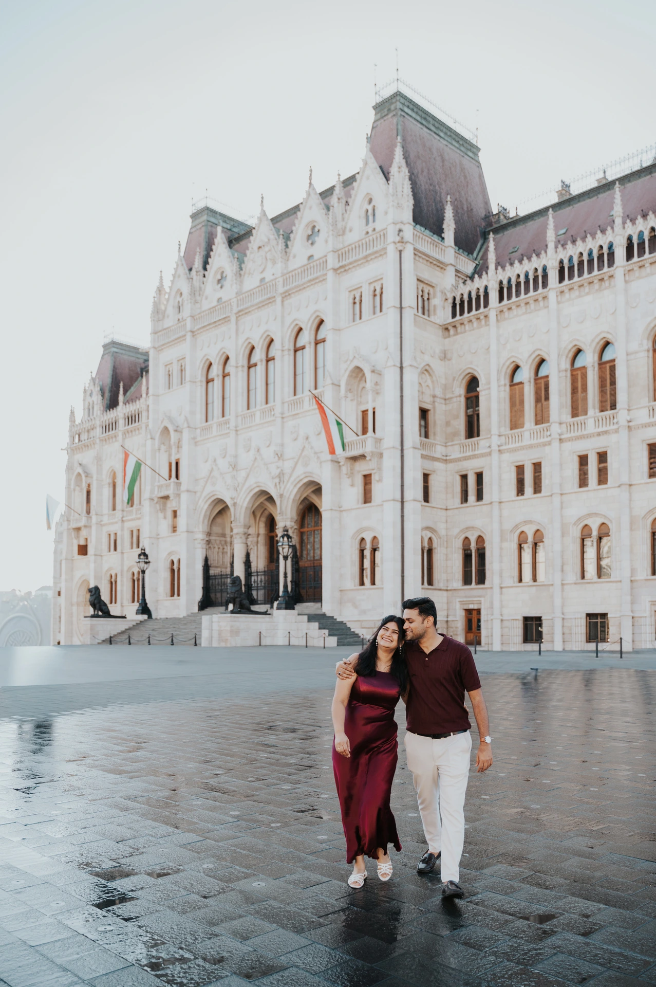 The couple walking together across the square beside Parliament’s pale stone façade during their couple photoshoot in Budapest.