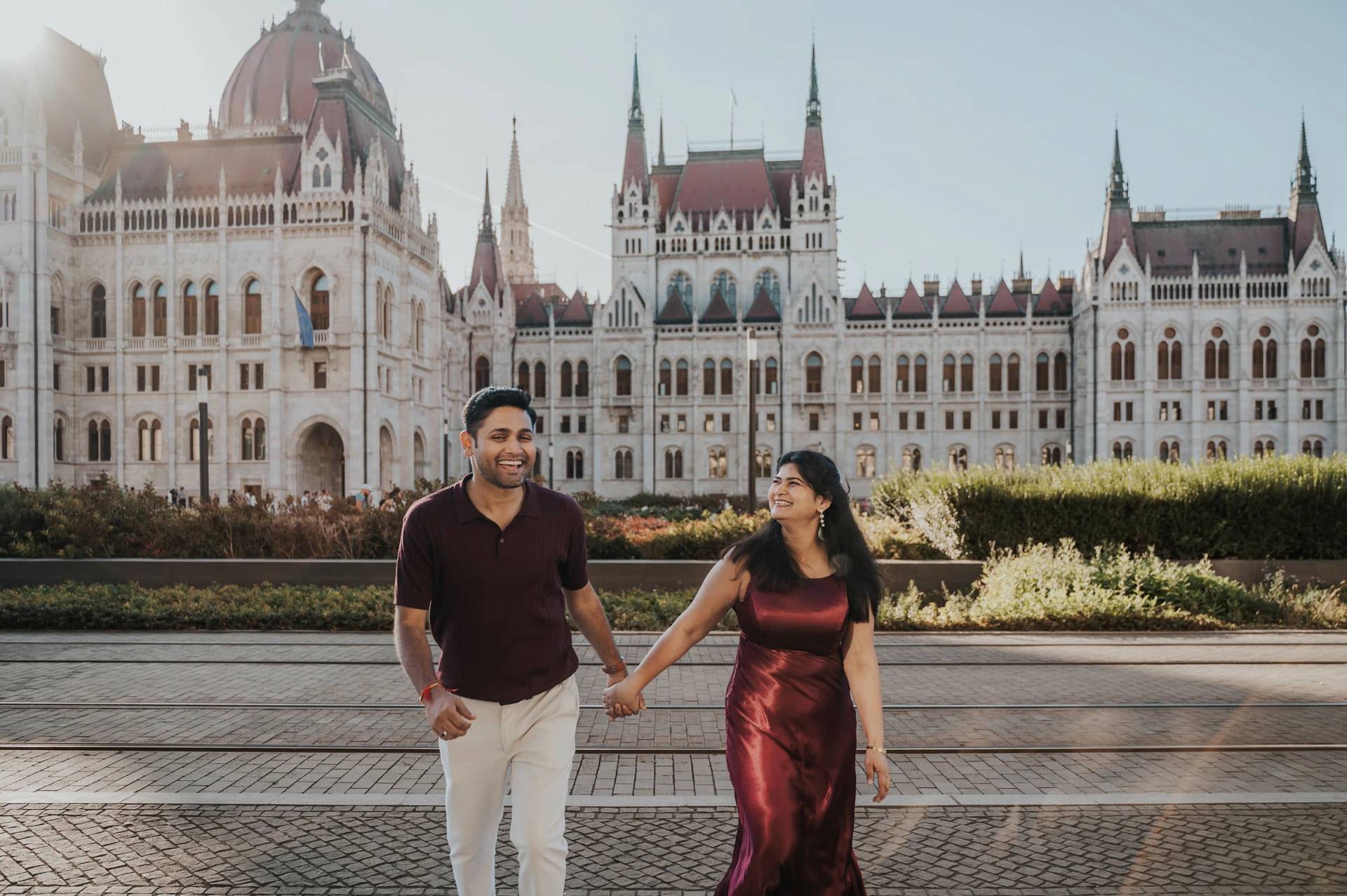 The couple smiling and holding hands in a wide portrait with Parliament’s pale façade behind them during their Budapest couple photoshoot.