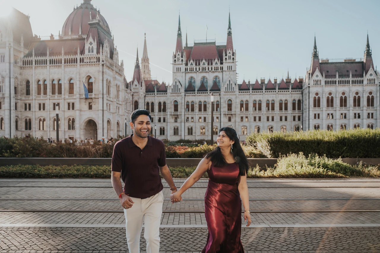A wide Parliament portrait at a Budapest couple photoshoot The couple smiling and holding hands in a wide portrait with Parliament’s pale façade behind them during their Budapest couple photoshoot.
