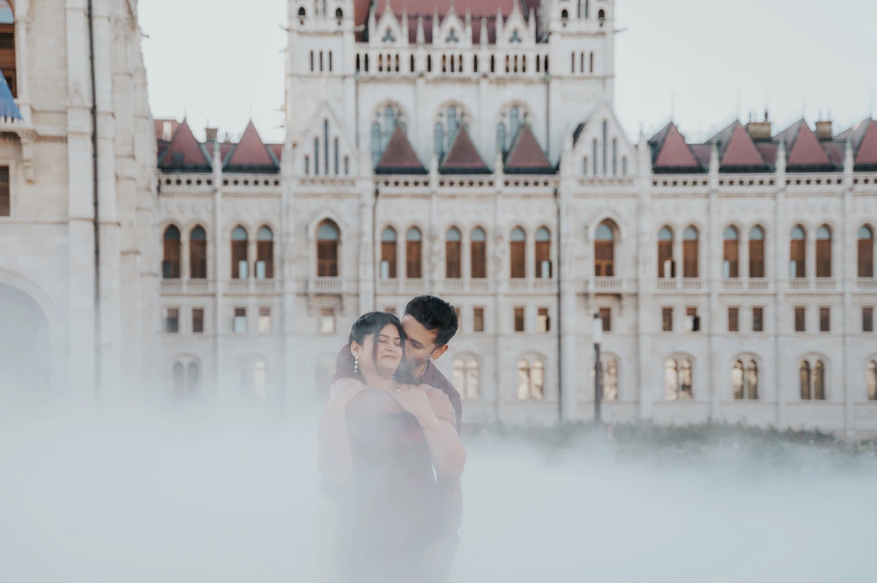A wide misty view at a Budapest couple photoshoot by Parliament A wide view of the couple embracing through drifting mist in front of Parliament’s ornate stone façade during their Budapest couple photoshoot.
