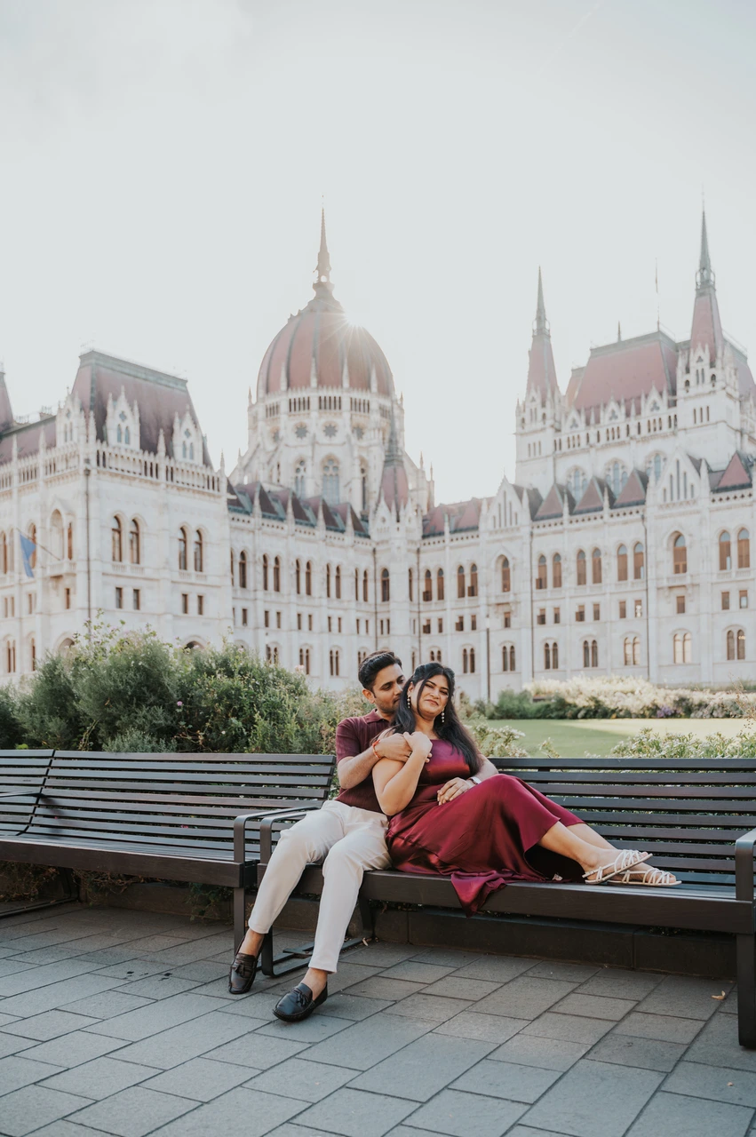 A playful bench portrait at a Budapest couple photoshoot The couple sharing a playful seated portrait on a wooden bench, with Parliament and the flowerbeds behind them, during their Budapest couple photoshoot.