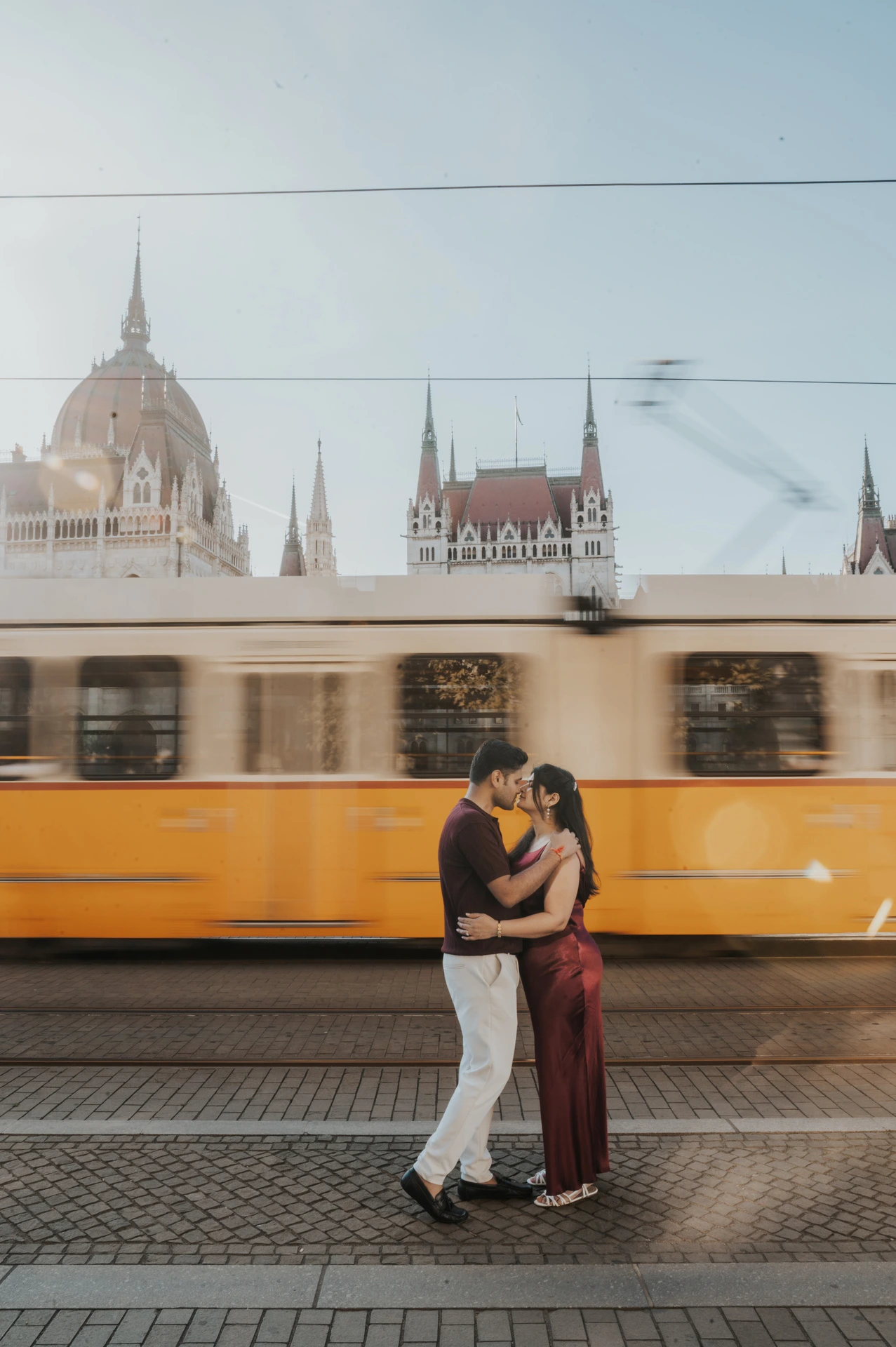The couple embracing beside the tracks as a yellow tram passes in front of Parliament during their Budapest couple photoshoot.