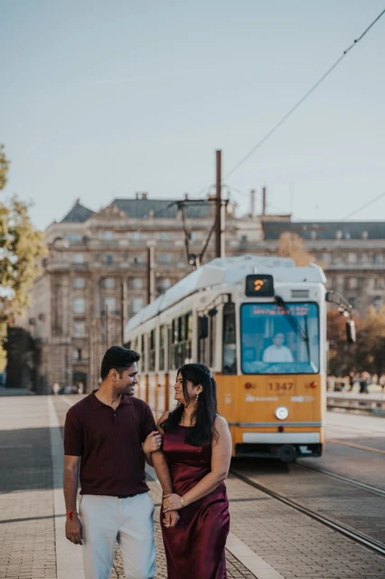 A Budapest couple photoshoot walk beside the yellow tram The couple walking together with a yellow tram and city buildings behind them during their couple photoshoot in Budapest.