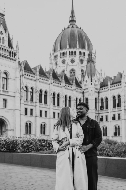 Budapest couple photoshoot: a black-and-white embrace beside Parliament Black-and-white view of the couple holding each other in front of Parliament, with the garden wall and flowers beside them during their Budapest couple photoshoot.