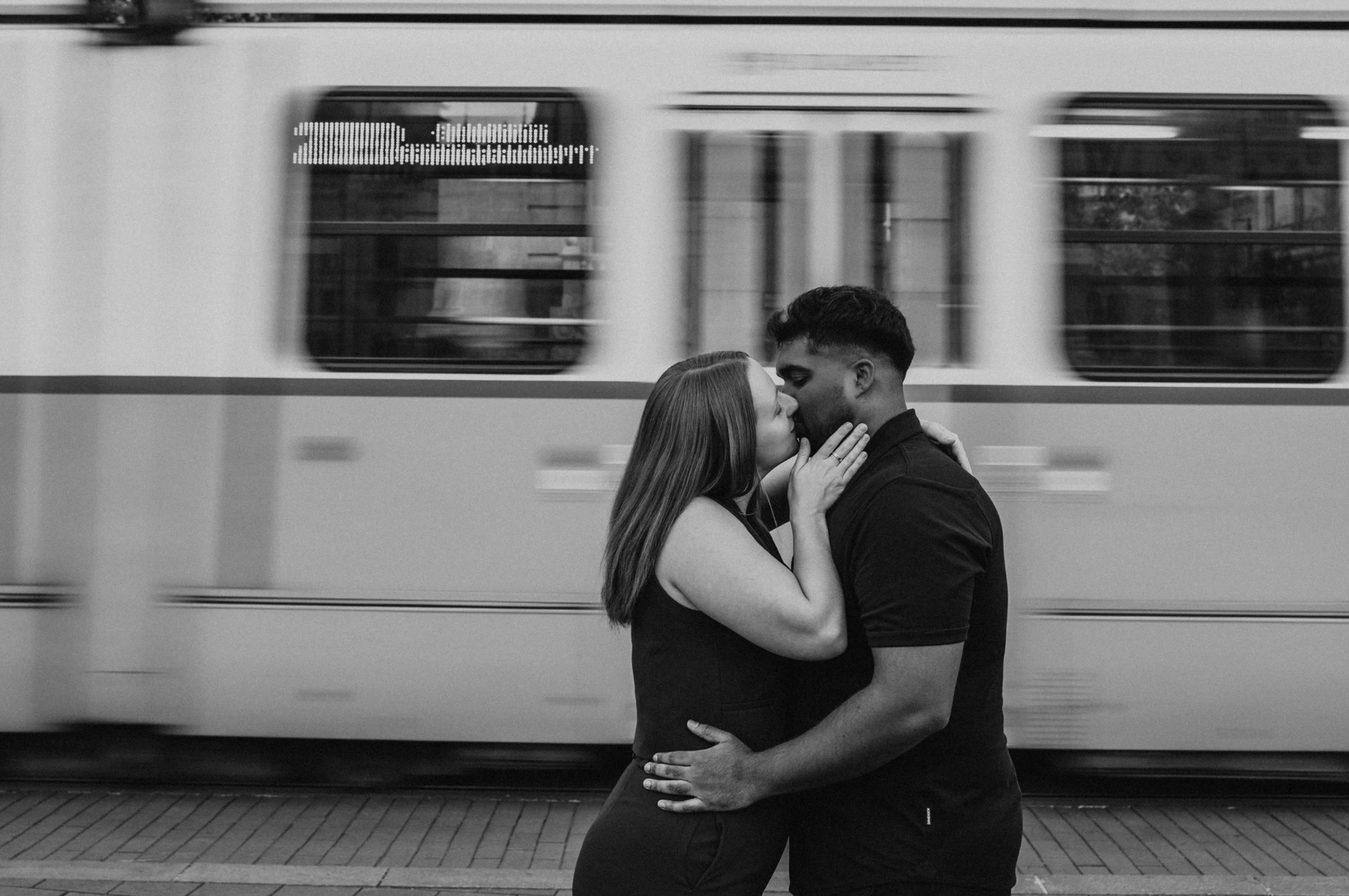 Black-and-white landscape photograph of the couple kissing as a Budapest tram passes behind them during their Budapest couple photoshoot.