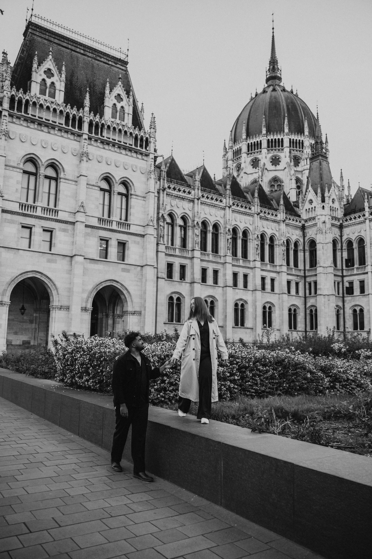 Black-and-white photograph of the couple holding hands beside the low garden wall, with her standing on the ledge and Parliament behind them during their Budapest couple photoshoot.