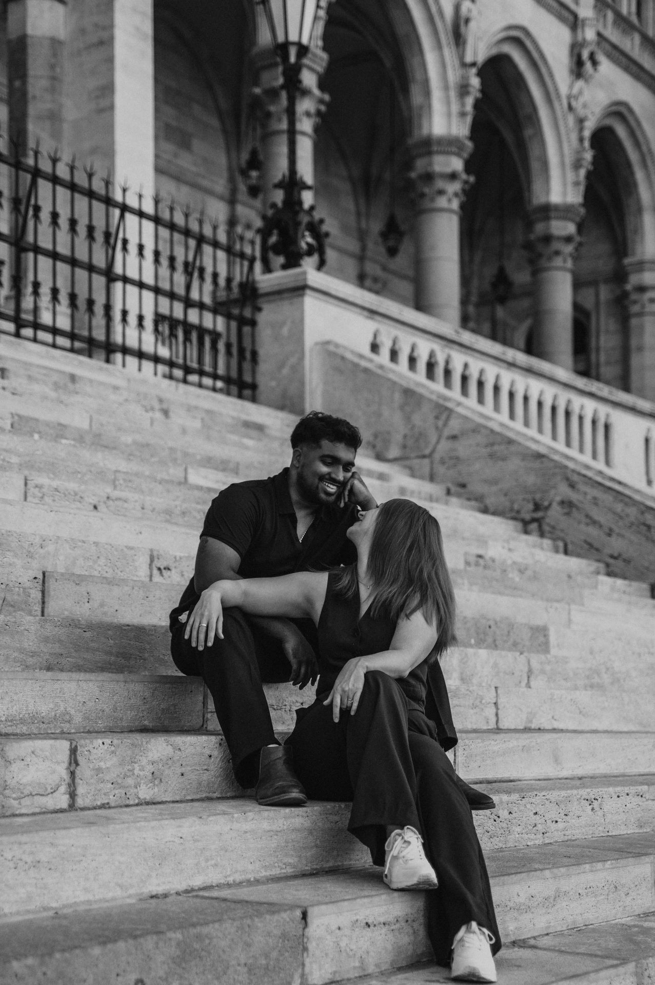 Black-and-white photograph of the couple seated together on Parliament’s steps, framed by the pale stonework and iron railings during their Budapest couple photoshoot.
