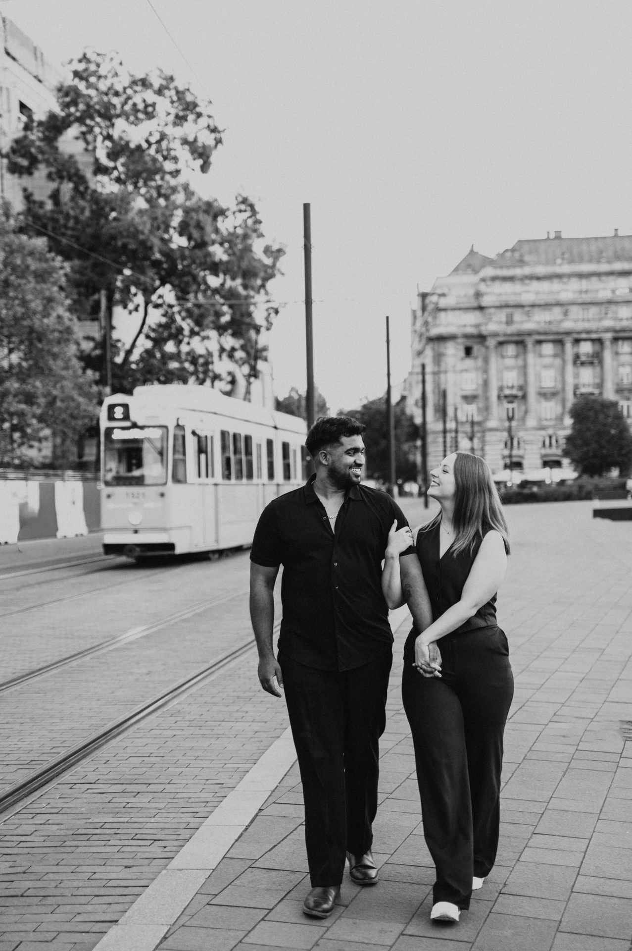Budapest couple photoshoot: a black-and-white walk beside the trams Black-and-white photograph of the couple walking hand in hand near the tram route, smiling towards each other during their Budapest couple photoshoot.