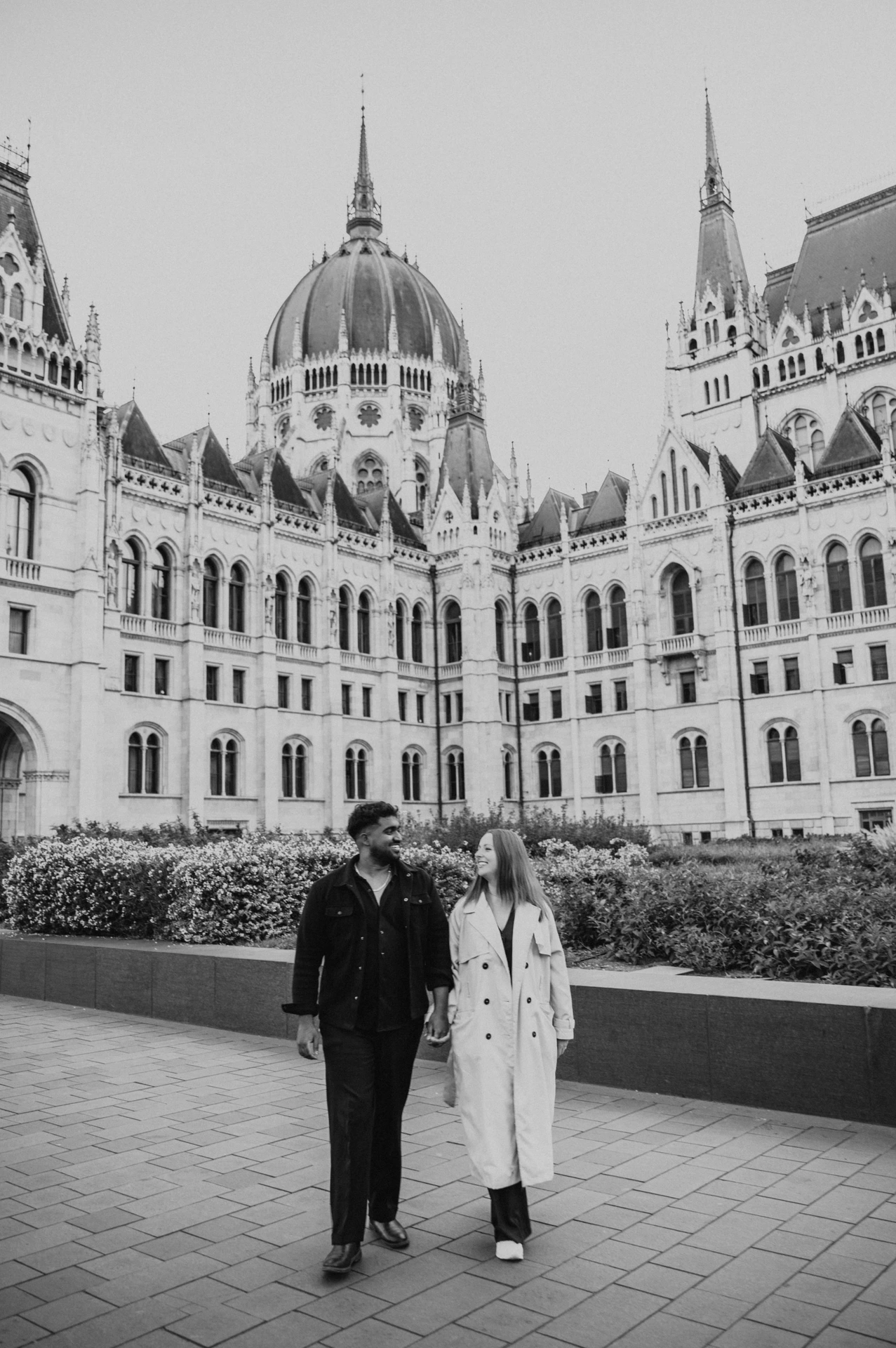 Black-and-white photograph of the couple walking side by side in front of the Hungarian Parliament and its flowerbeds during their Budapest couple photoshoot.