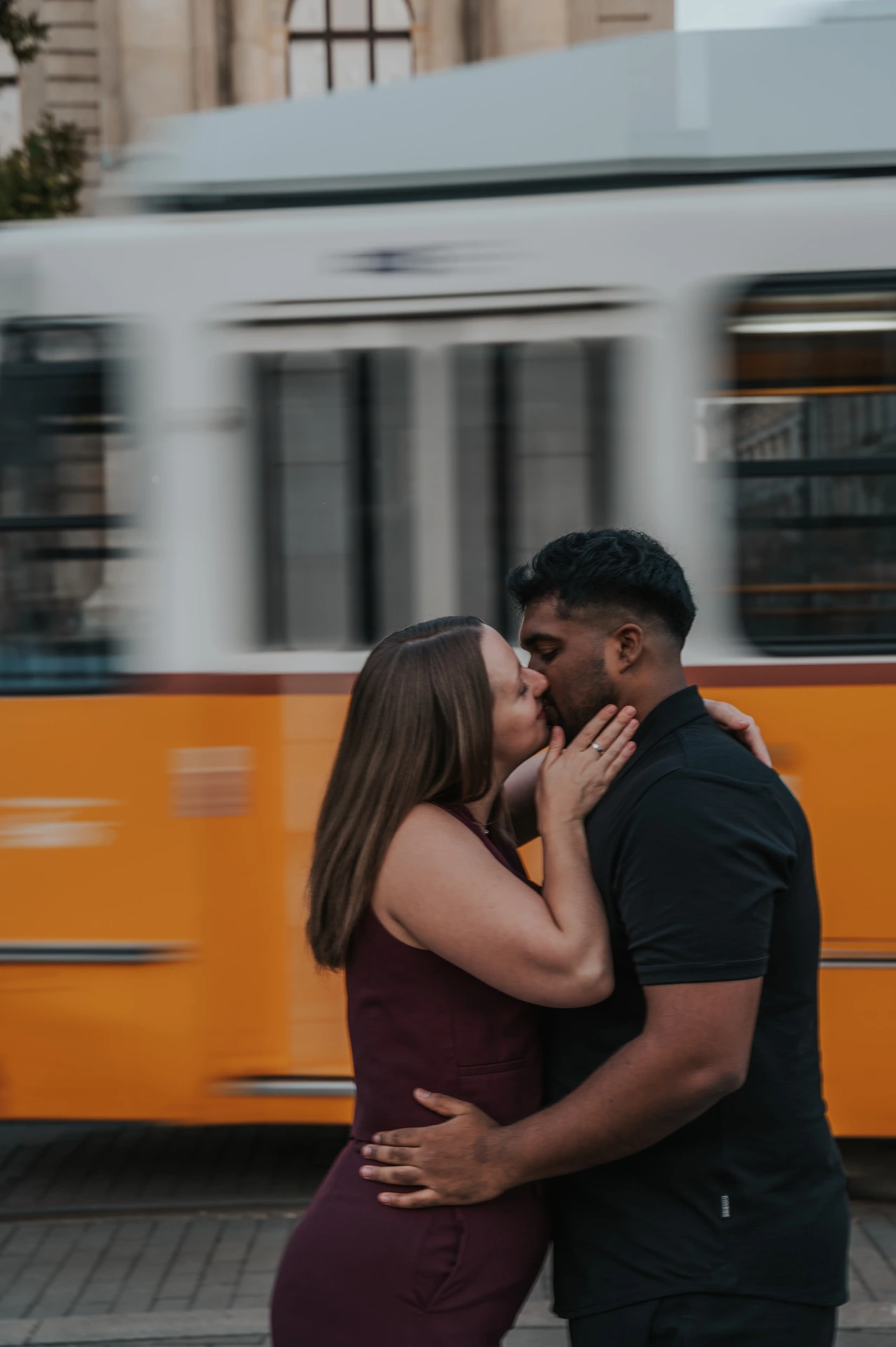 The couple kissing closely in front of the yellow tram, with his arm around her waist and her hand on his cheek during their Budapest couple photoshoot.