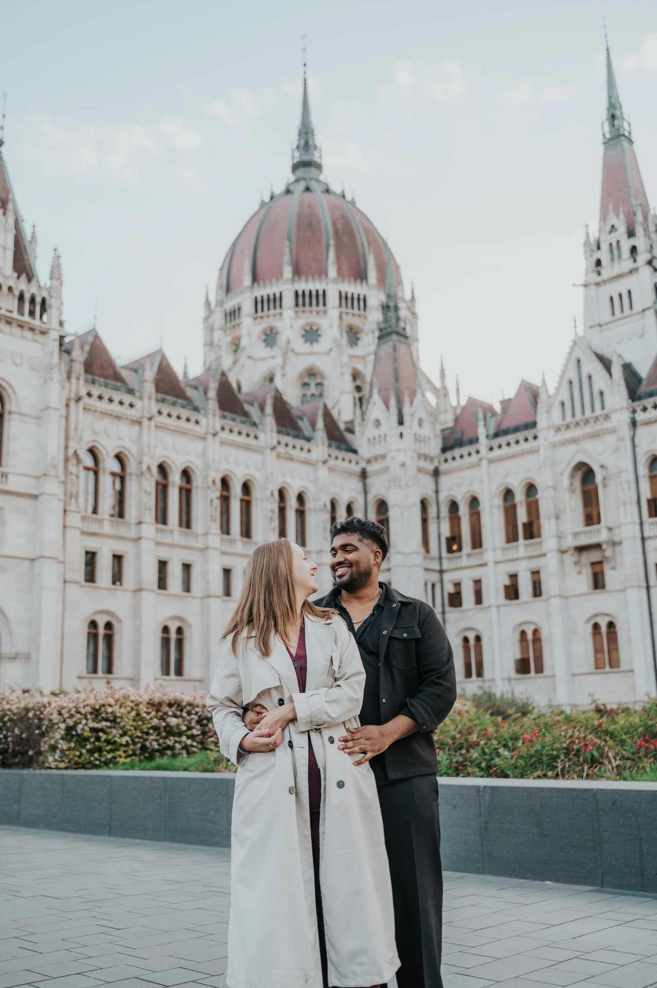 Budapest couple photoshoot: a closer portrait in front of Parliament A closer view of the couple smiling together beside the Parliament gardens, with the ornate façade rising behind them during their Budapest couple photoshoot.
