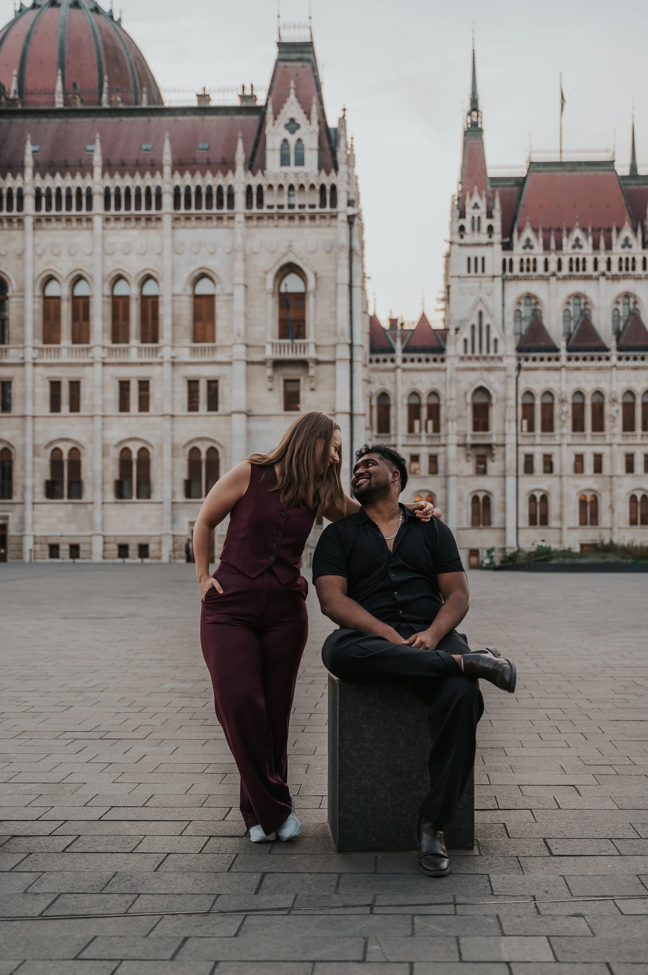 Budapest couple photoshoot: a closer portrait together in Parliament Square A closer view of the couple together beside a stone bollard, with her standing behind him and Parliament in the backdrop during their Budapest couple photoshoot.