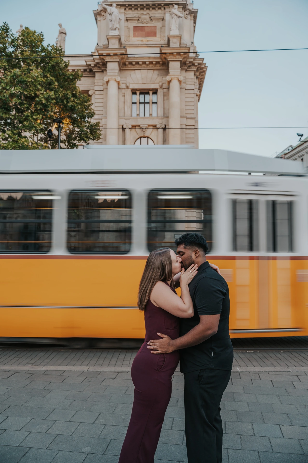 A full-length view of the couple kissing beside the tram route, with the yellow tram and a historic Budapest building behind them during their Budapest couple photoshoot.