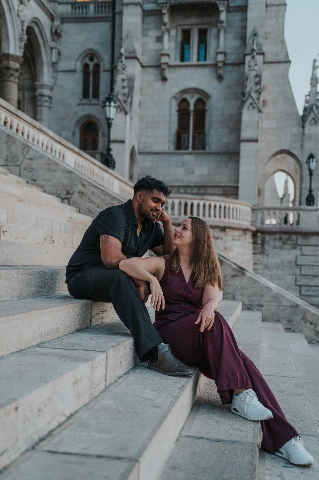 The couple kissing while seated close together on the stone staircase beside Parliament’s ornate riverside façade during their Budapest couple photoshoot.