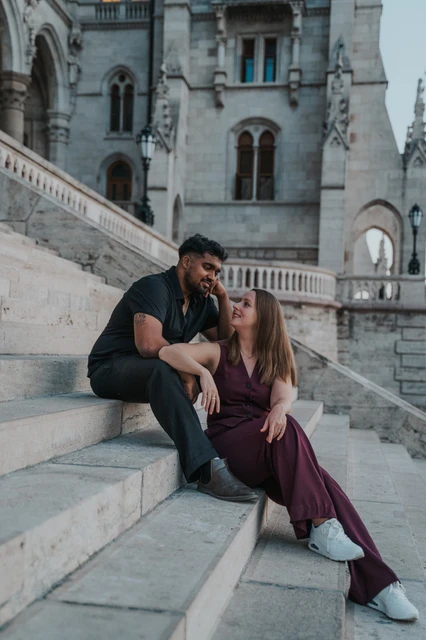Budapest couple photoshoot: a kiss beneath Parliament’s carved arches The couple kissing while seated close together on the stone staircase beside Parliament’s ornate riverside façade during their Budapest couple photoshoot.