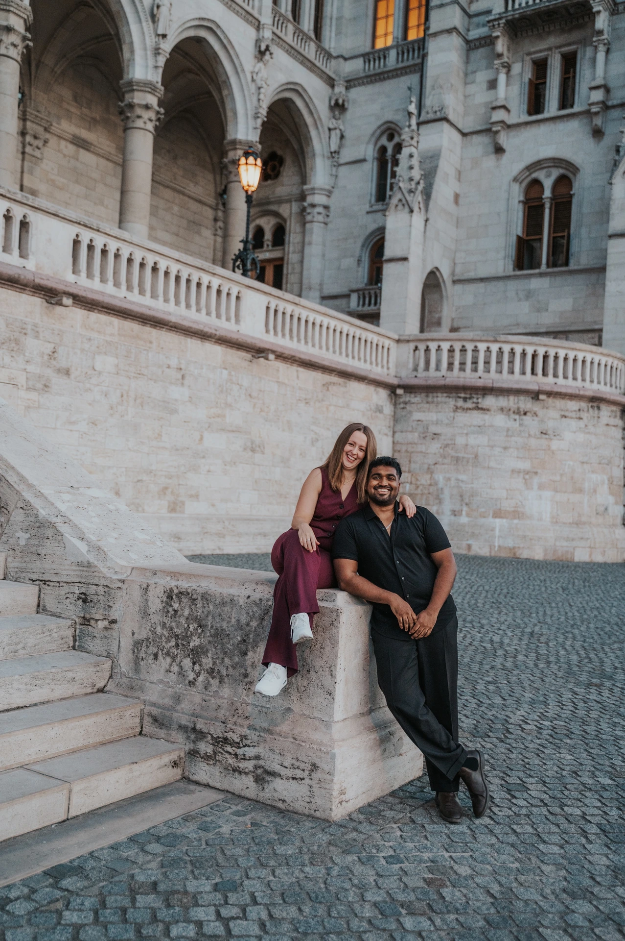 The couple kissing beside the low stone pillar at Parliament’s staircase, with the ornate façade above them during their Budapest couple photoshoot.