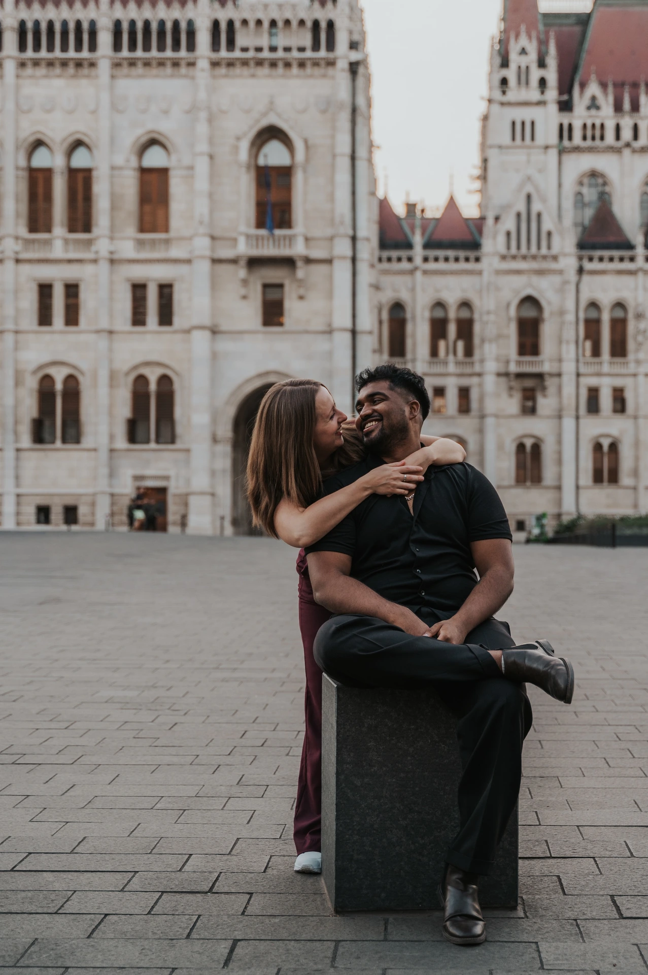 She leans in to kiss him while he sits on a stone bollard, with Parliament’s pale façade behind them during their Budapest couple photoshoot.