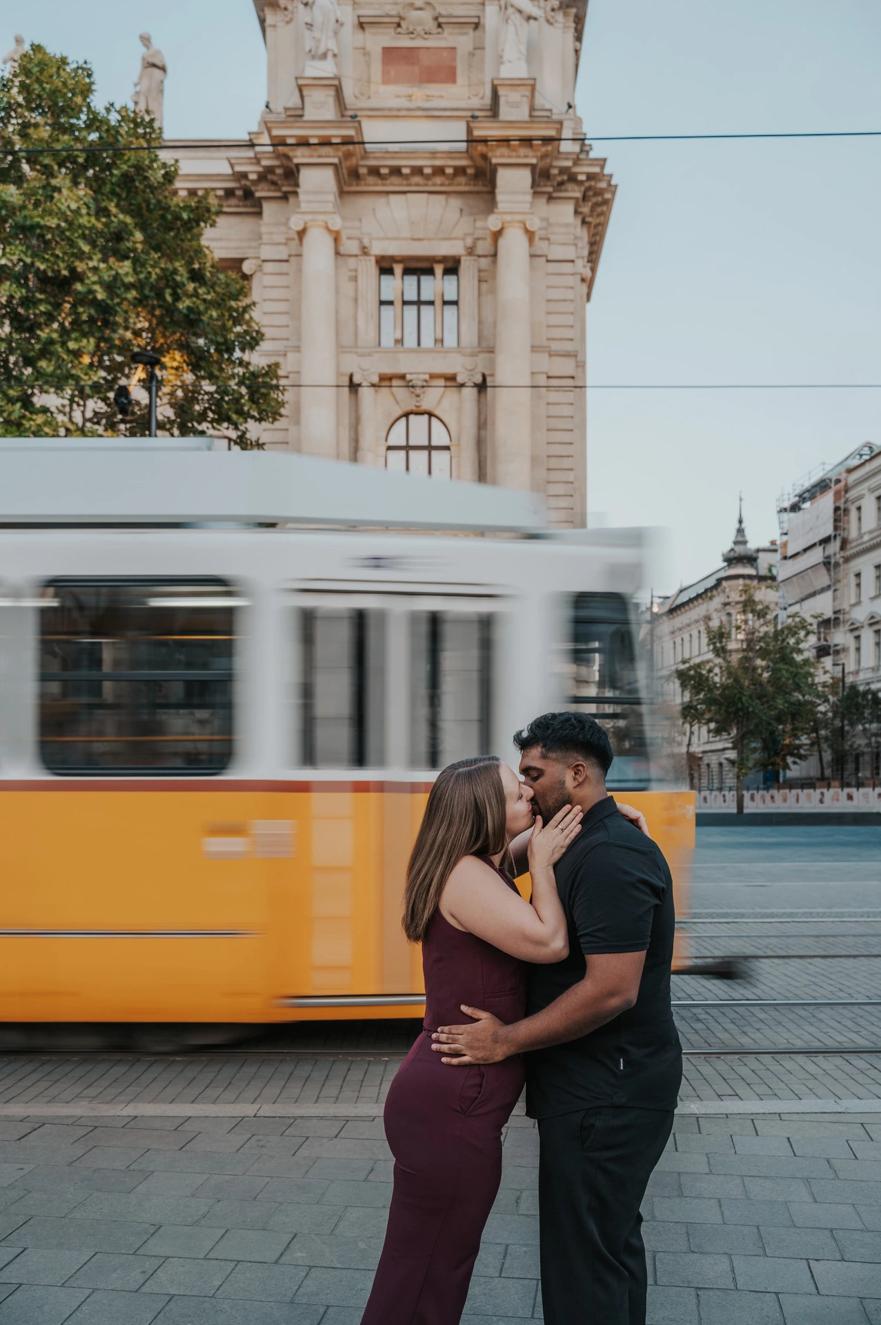Budapest couple photoshoot: a kiss in front of a passing yellow tram The couple kissing in an embrace as a yellow Budapest tram passes behind them during their Budapest couple photoshoot.