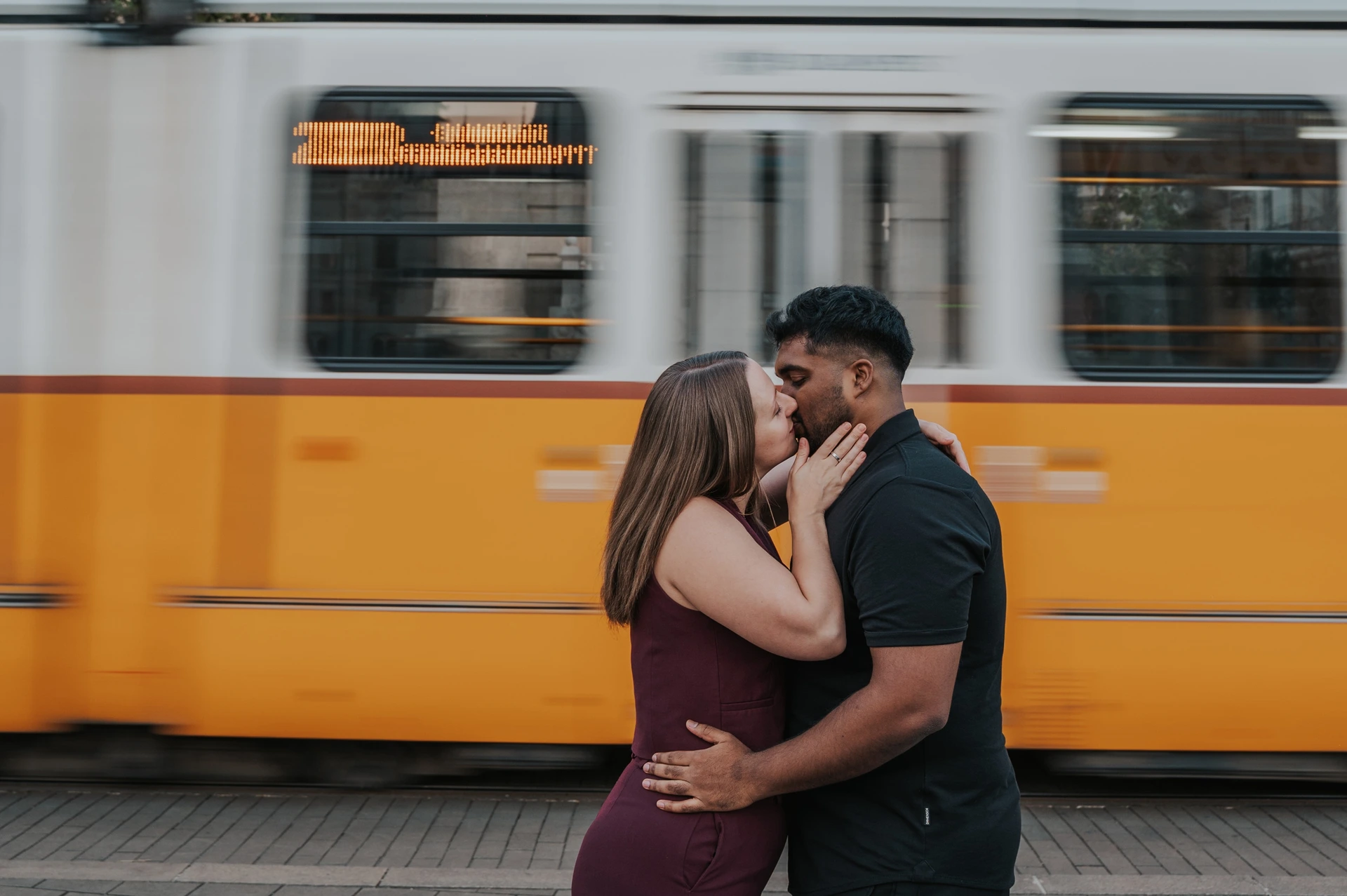 Budapest couple photoshoot: a landscape kiss with a yellow tram backdrop A landscape photograph of the couple kissing, her hand against his cheek, with the moving yellow Budapest tram filling the background during their Budapest couple photoshoot.
