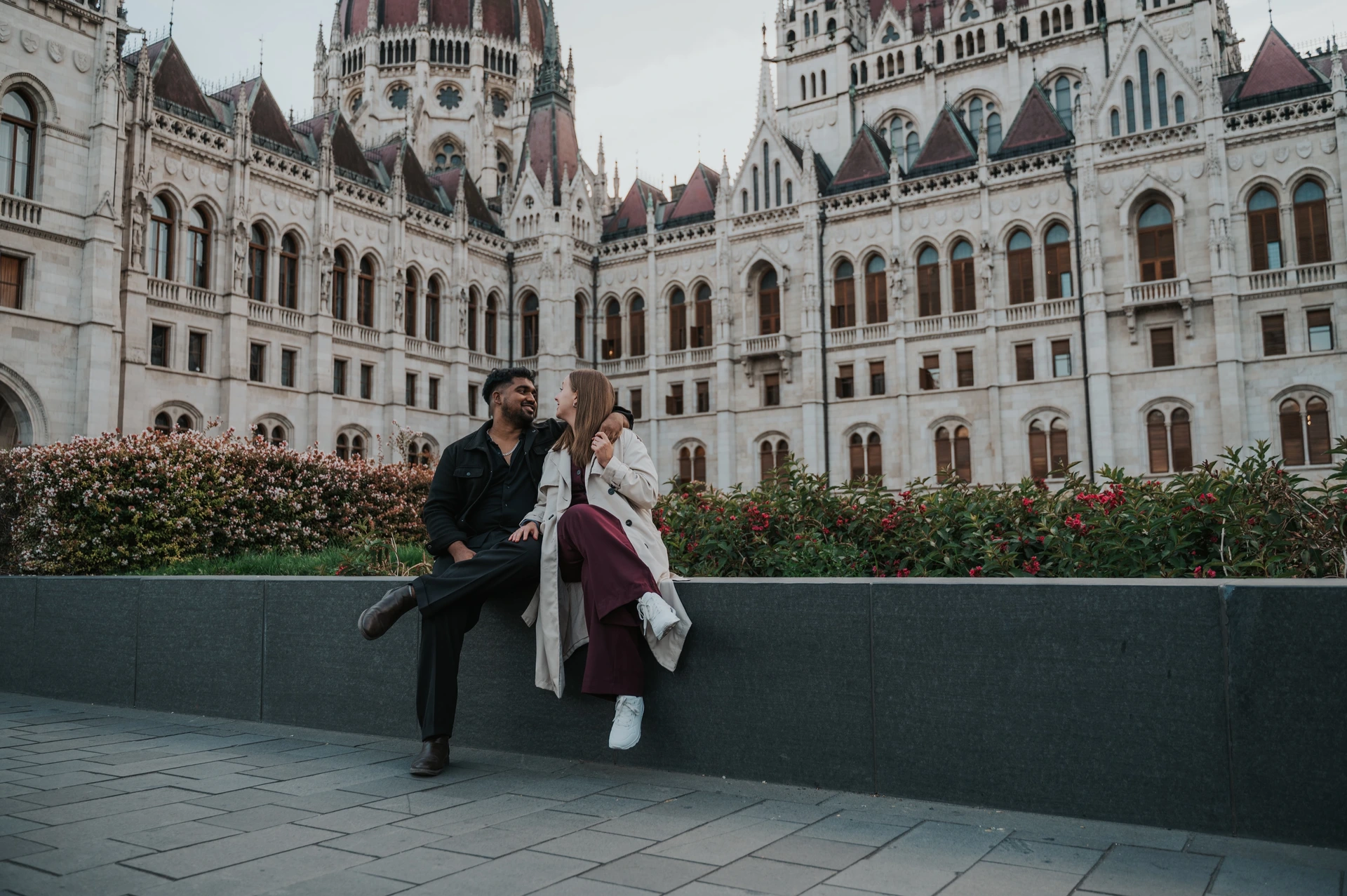 Budapest couple photoshoot: a landscape portrait with Parliament’s façade A landscape view of the couple sitting together on the garden wall, with Parliament’s ornate façade and flowerbeds behind them during their Budapest couple photoshoot.