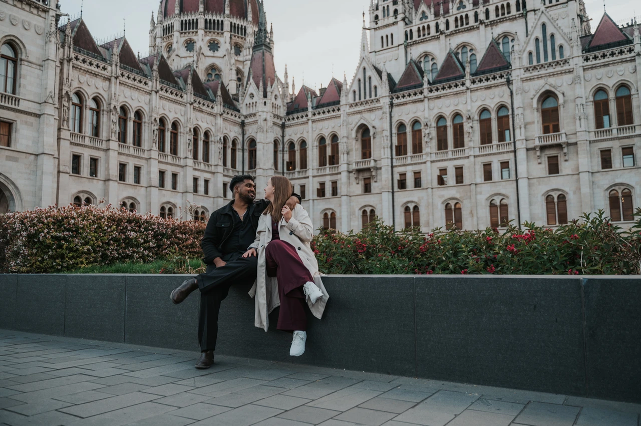 Budapest couple photoshoot: a landscape portrait with Parliament’s façade A landscape view of the couple sitting together on the garden wall, with Parliament’s ornate façade and flowerbeds behind them during their Budapest couple photoshoot.