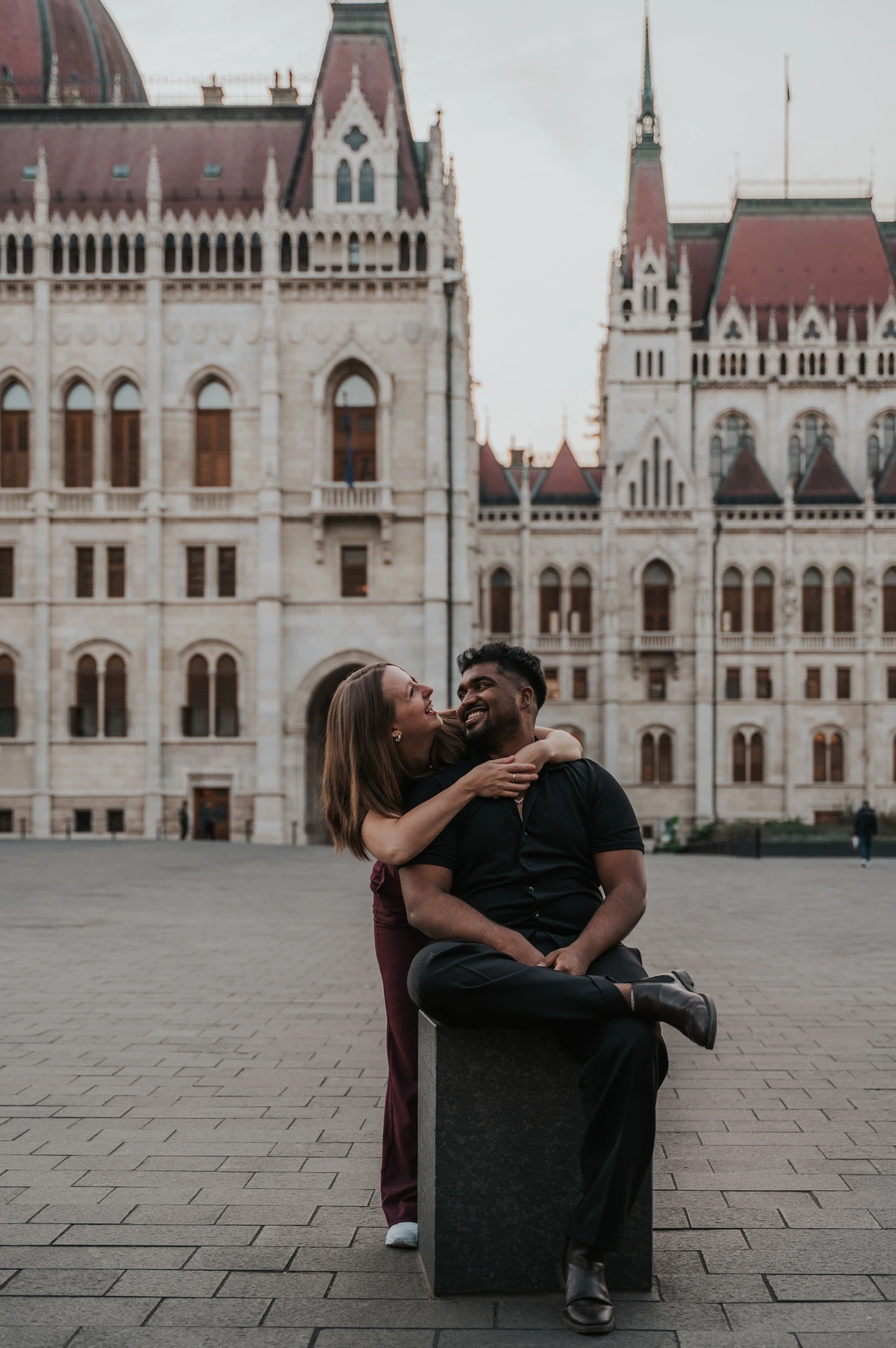 The couple kissing and embracing beside the stone bollard in the open square outside the Hungarian Parliament during their Budapest couple photoshoot.
