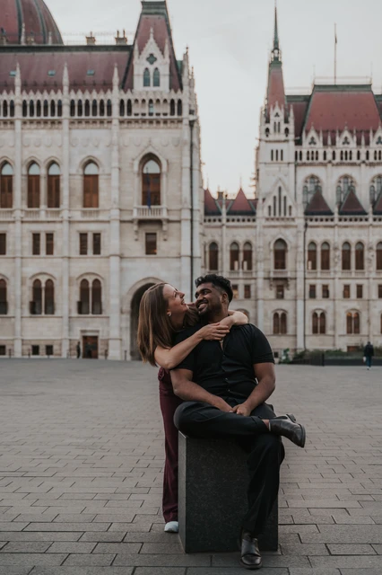 Budapest couple photoshoot: a playful embrace in the square The couple kissing and embracing beside the stone bollard in the open square outside the Hungarian Parliament during their Budapest couple photoshoot.