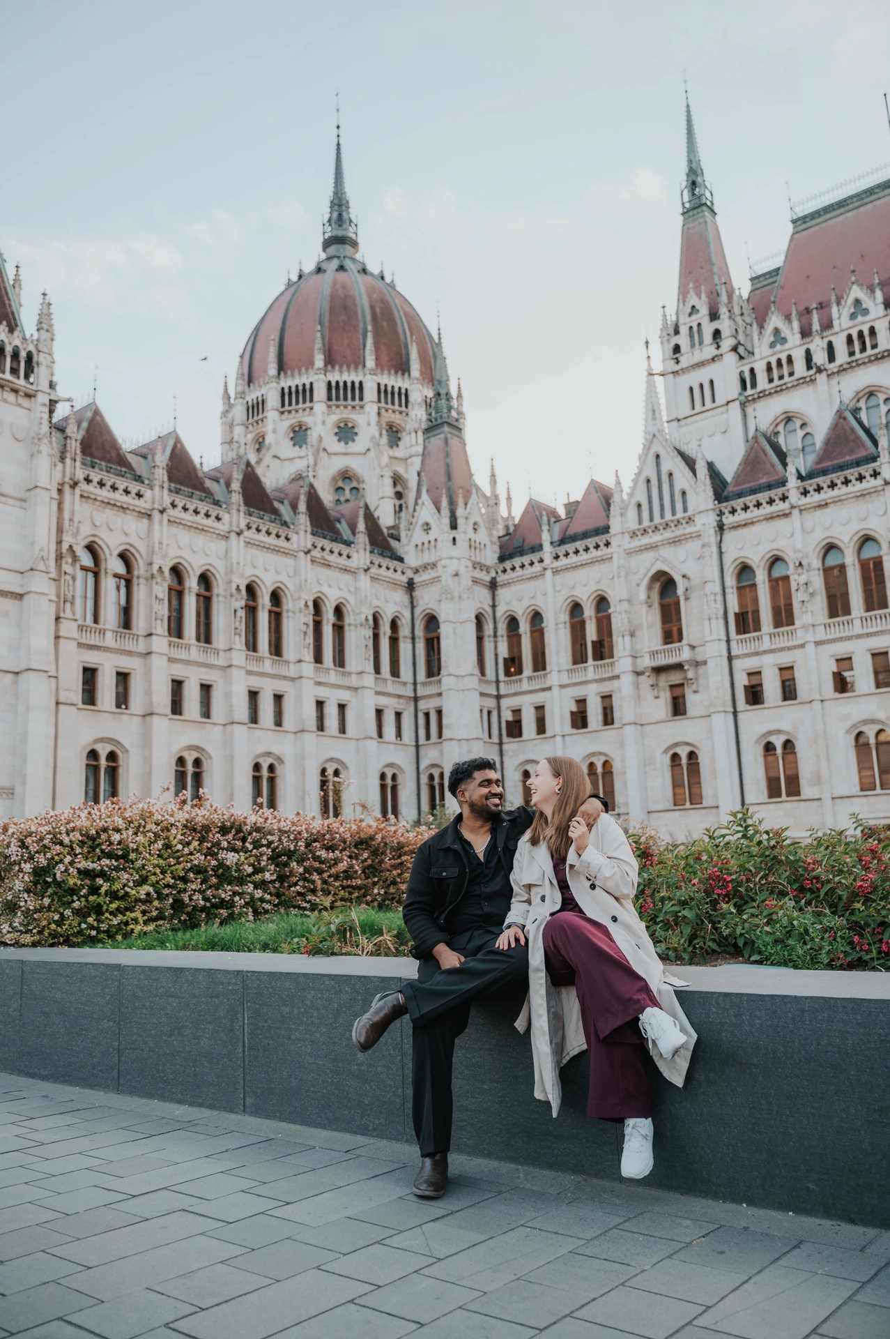 The couple sharing a playful moment while sitting on the garden wall, with the full height of Parliament’s dome above them during their Budapest couple photoshoot.