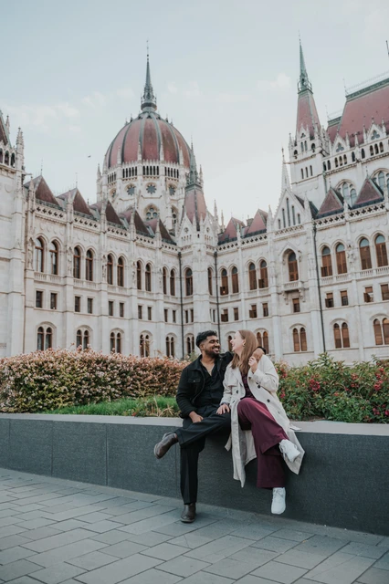 Budapest couple photoshoot: a playful moment beneath Parliament’s dome The couple sharing a playful moment while sitting on the garden wall, with the full height of Parliament’s dome above them during their Budapest couple photoshoot.