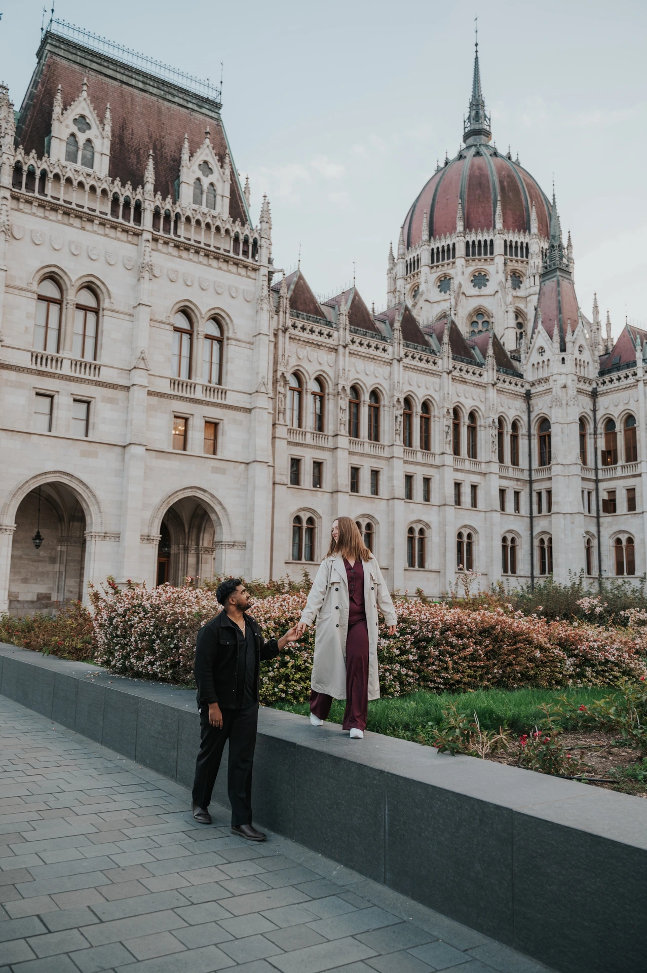 A wide view of her walking along the garden wall as he holds her hand from the paving below, with Parliament above them during their Budapest couple photoshoot.