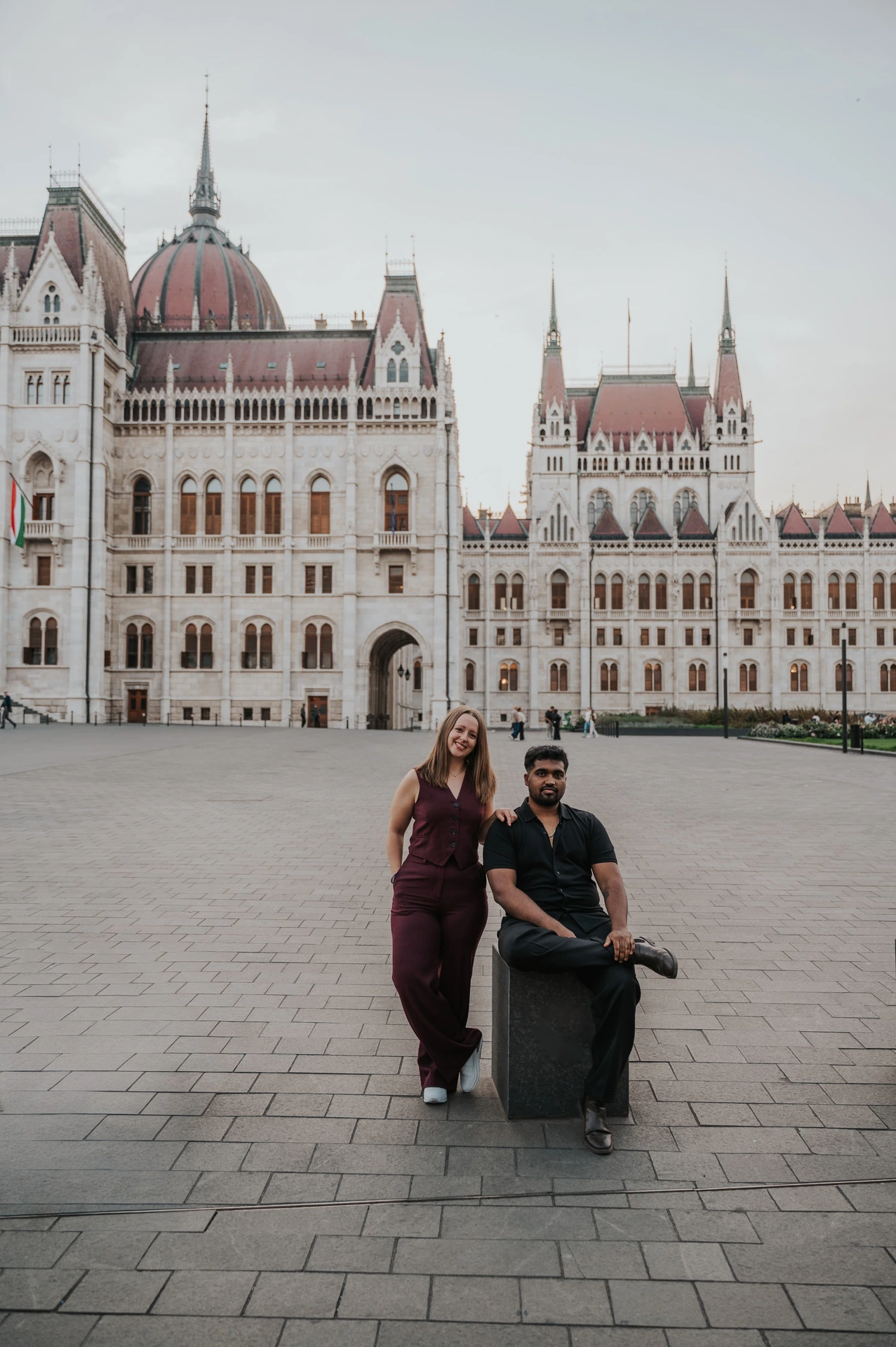 He sits on a low stone bollard while she stands close behind him, with the Hungarian Parliament across the square during their Budapest couple photoshoot.