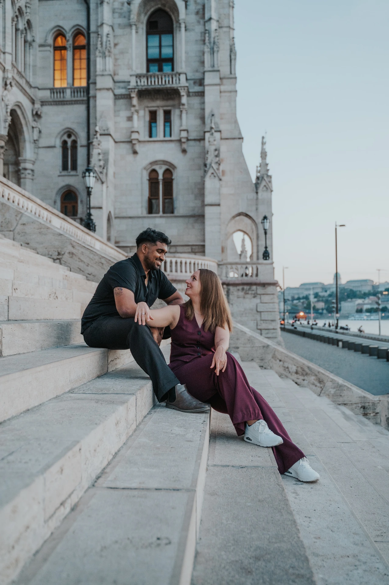 Budapest couple photoshoot: a quiet shared look on the riverside steps The couple seated close together on Parliament’s riverside staircase, looking towards each other beneath the carved arches during their Budapest couple photoshoot.