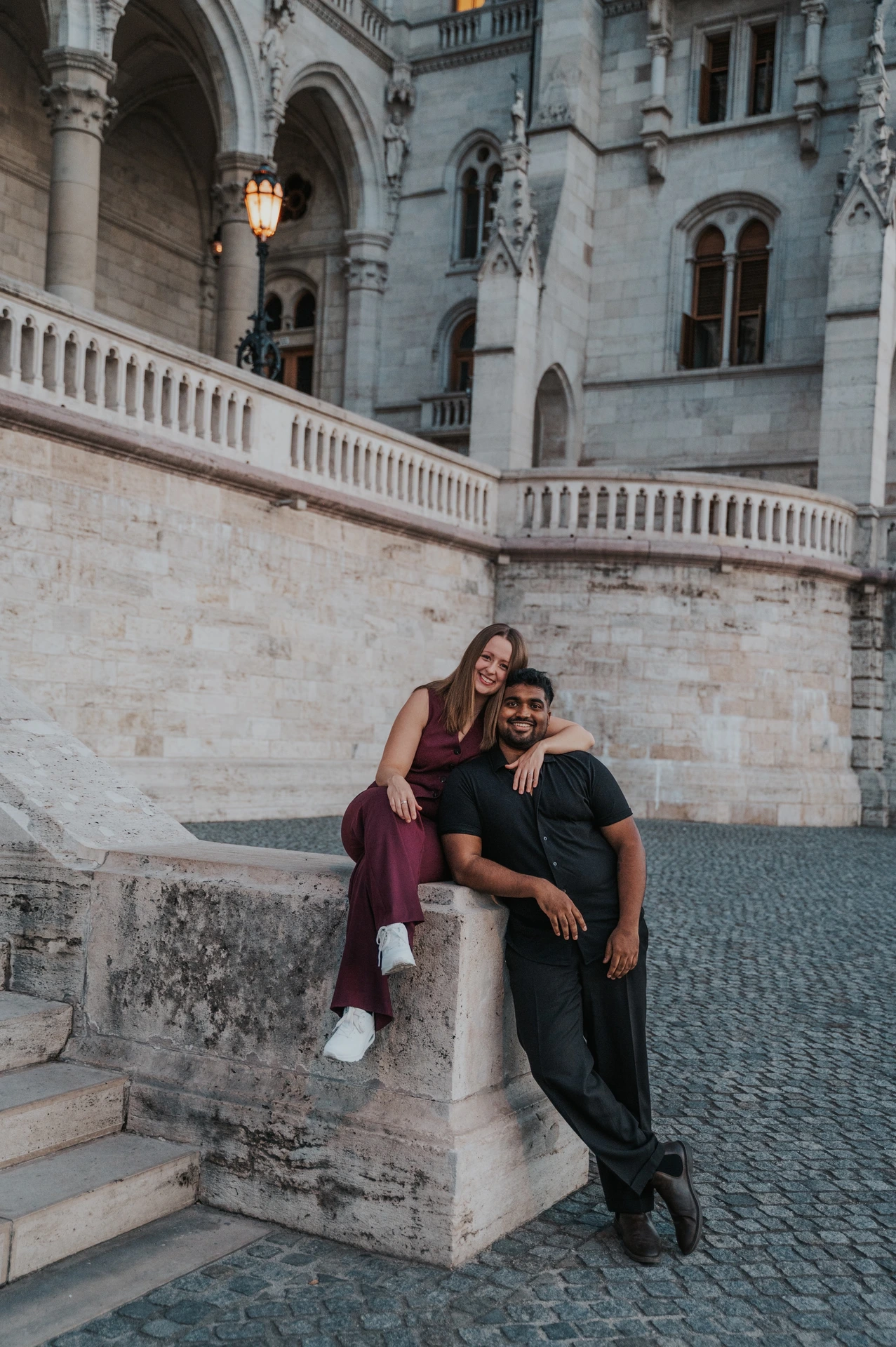 The couple together by the stone pillar beside Parliament’s steps, with her seated and him leaning close beneath the arches during their Budapest couple photoshoot.