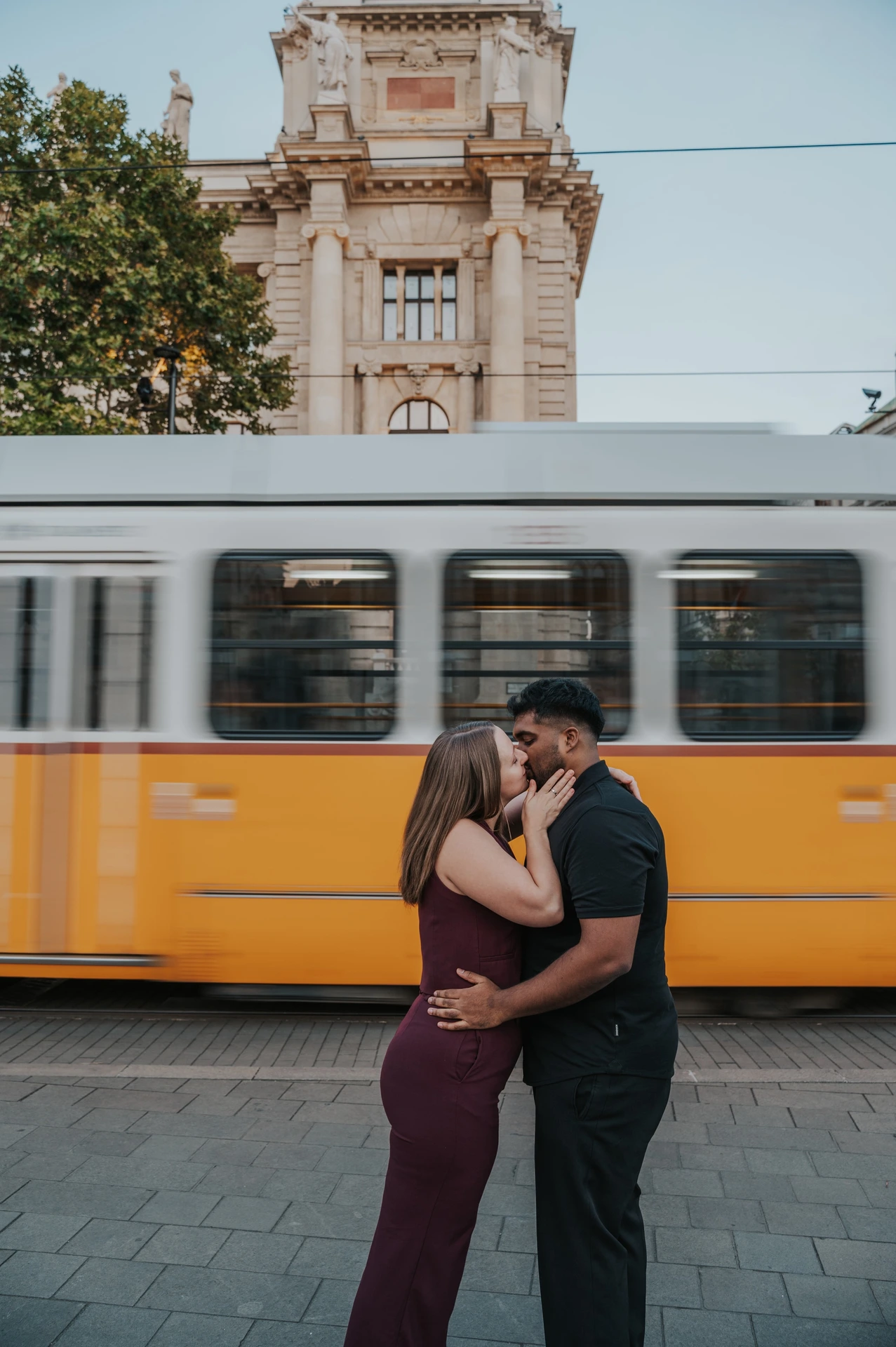 Budapest couple photoshoot: a shared kiss against the passing tram The couple holding each other in a kiss as the yellow-and-white Budapest tram moves through the background during their Budapest couple photoshoot.