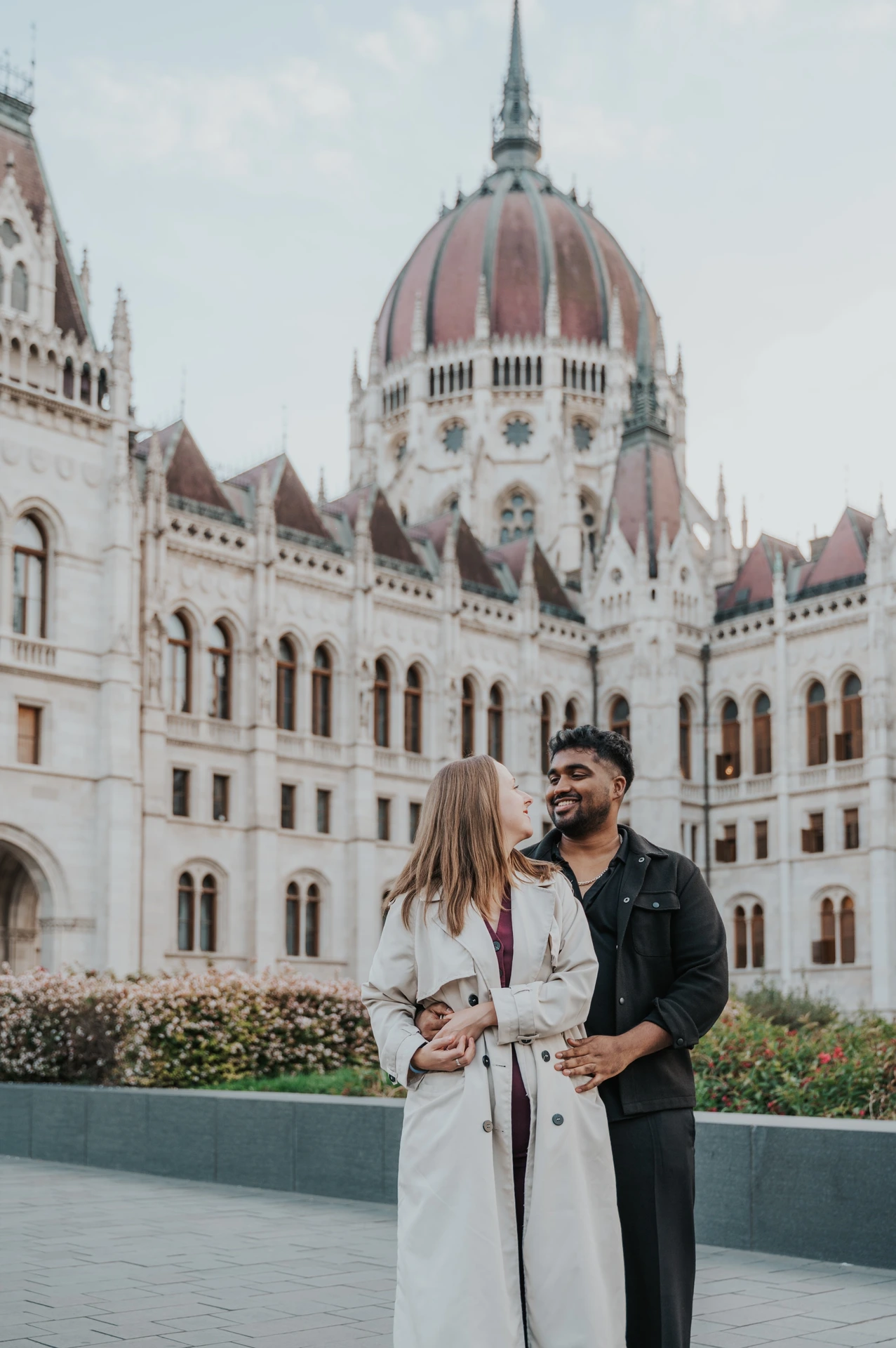 The couple looking at each other in an embrace beside the flowerbeds, with the Hungarian Parliament in the background during their Budapest couple photoshoot.
