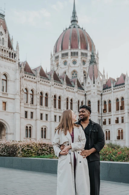 Budapest couple photoshoot: a shared look below Parliament’s dome The couple looking at each other in an embrace beside the flowerbeds, with the Hungarian Parliament in the background during their Budapest couple photoshoot.