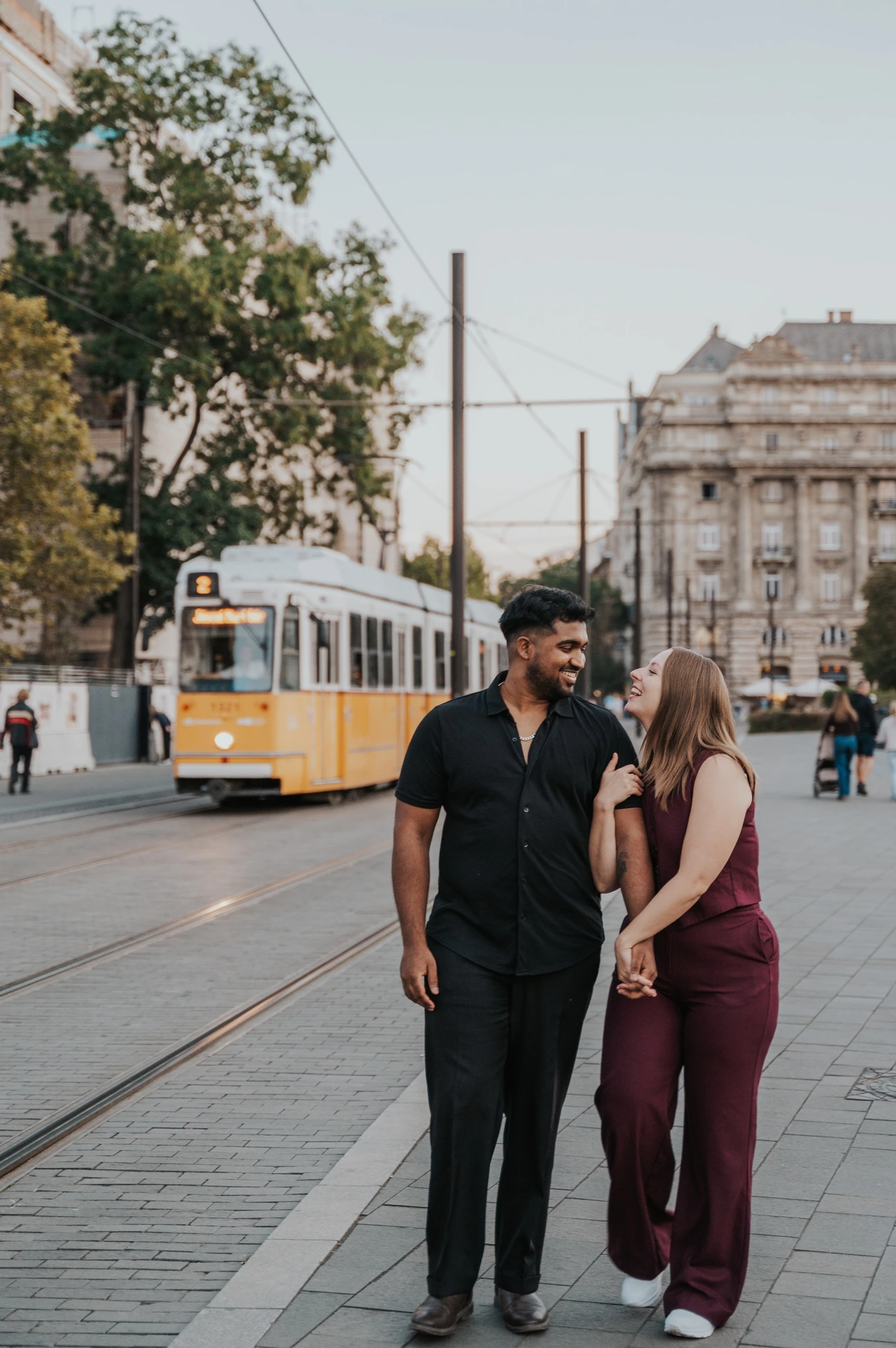 The couple looking at one another as they walk together beside the tram route, with a yellow tram in the background during their Budapest couple photoshoot.