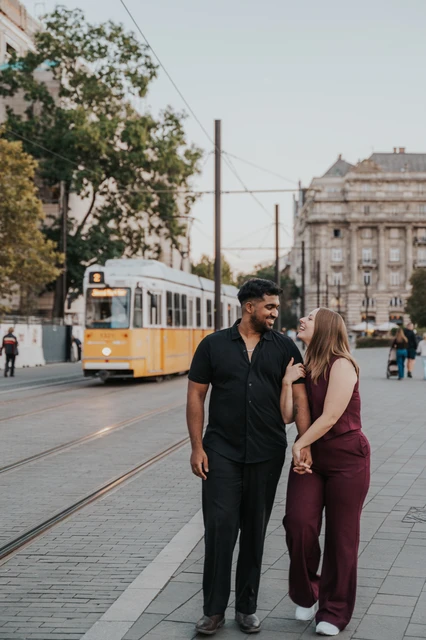 Budapest couple photoshoot: a shared smile on their Budapest walk The couple looking at one another as they walk together beside the tram route, with a yellow tram in the background during their Budapest couple photoshoot.