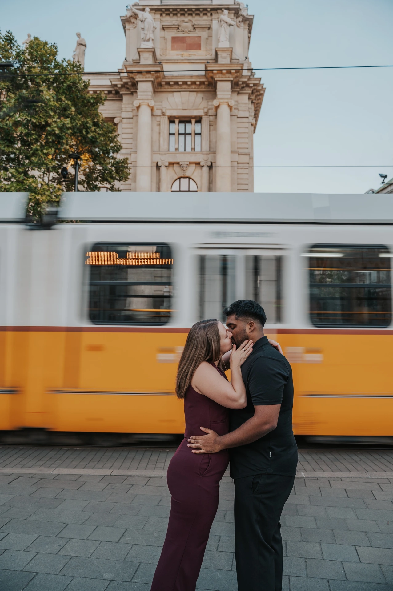 Budapest couple photoshoot: a wide city view and a tram-side kiss A wide vertical view of the couple kissing beside the yellow tram, with a tree and ornate city building above it during their Budapest couple photoshoot.