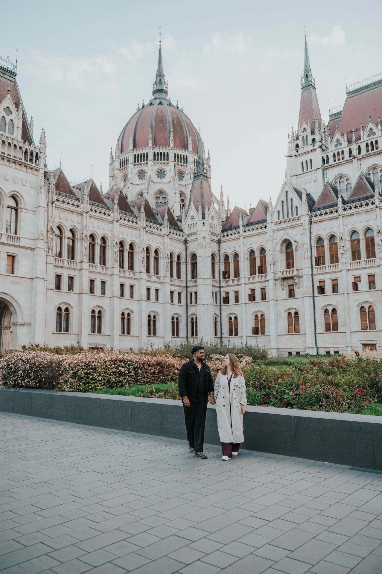 Budapest couple photoshoot: a wide portrait with Parliament in the backdrop The couple standing together beside the garden wall, with the Hungarian Parliament’s dome and tall stone façade above them during their Budapest couple photoshoot.