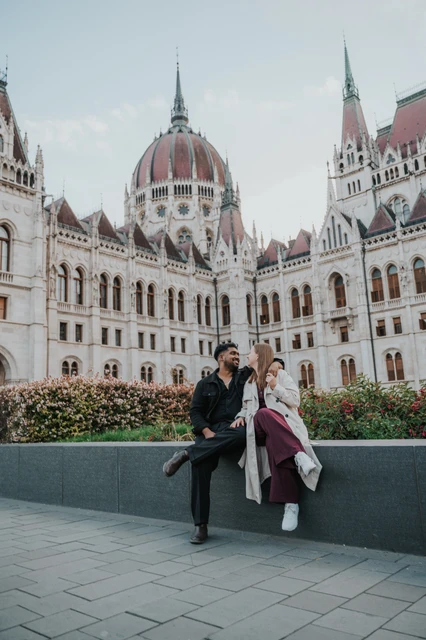 Budapest couple photoshoot: a wide seated portrait outside Parliament A wide vertical view of the couple seated on the garden ledge, framed by Parliament’s dome and stone towers during their Budapest couple photoshoot.