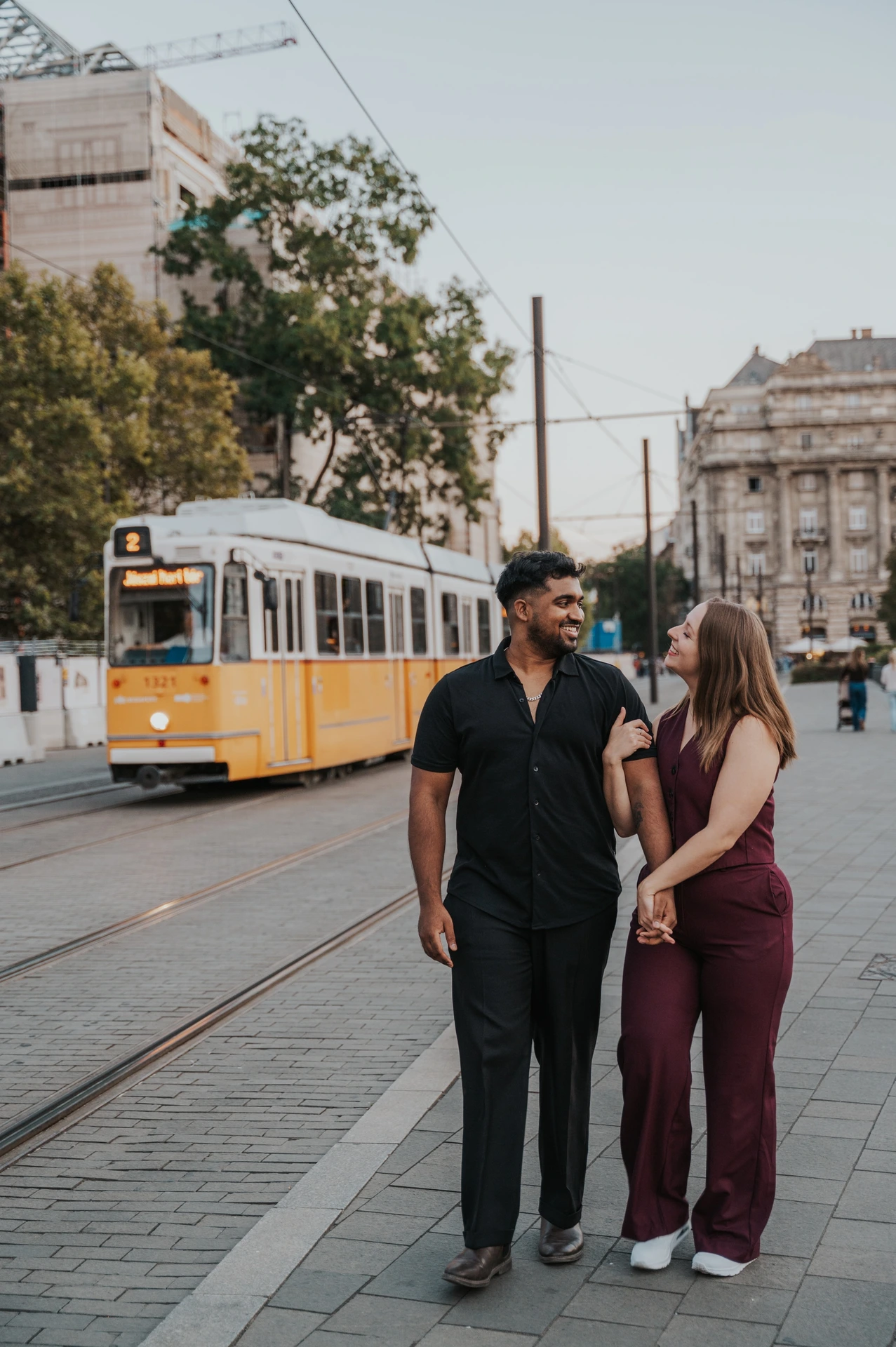 Budapest couple photoshoot: a wider portrait along the tram route A wide vertical view of the couple walking together beside the tram tracks, with a yellow tram and trees behind them during their Budapest couple photoshoot.