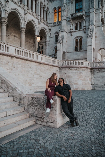 Budapest couple photoshoot: a wider staircase portrait at Parliament A wide vertical view of the couple beside the staircase’s stone pillar, framed by Parliament’s carved stonework and cobbled paving during their Budapest couple photoshoot.